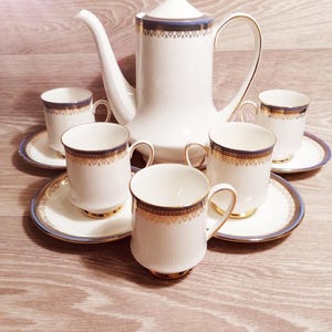 Puede incluir: Un juego de té de porcelana blanca con ribetes dorados y detalles azules. El juego incluye una tetera, platillos y tazas. Las tazas y la tetera tienen una banda decorativa dorada y azul alrededor del borde. El juego está dispuesto sobre una superficie de madera.