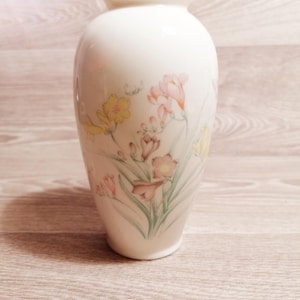 Puede incluir: Un jarrón de cerámica color crema con un diseño floral con flores amarillas, rosas y marrones con tallos y hojas verdes. El jarrón tiene un borde dorado y una forma redondeada.