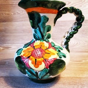 Puede incluir: Jarra de cerámica pintada a mano con un diseño floral. La jarra presenta una base blanca con una gran flor en tonos amarillos, naranjas y rosas. El asa es un diseño de cuerda trenzada en verde y blanco. El borde y la base son verdes.