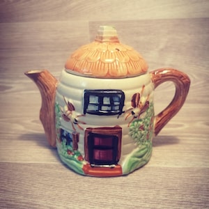 Pode incluir: Uma chaleira de cerâmica em forma de casa. A chaleira apresenta uma tampa em forma de telhado de colmo, uma pega e um bico castanhos e uma fachada pintada com uma porta, janelas e detalhes florais. A paleta de cores inclui creme, castanho, verde e vermelho.
