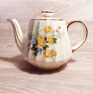 Könnte beinhalten: Cremefarbene Vintage-Teekanne mit floralem Design mit gelben Rosen und blauen Akzenten. Die Teekanne hat einen goldenen Besatz an Griff, Ausguss und Deckel. Die Teekanne steht auf einer hellen Holzoberfläche.