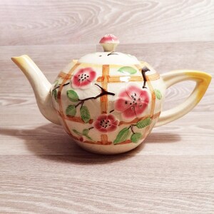 May include: Cream-colored ceramic teapot with a floral design. The teapot features pink flowers, green leaves, and a brown lattice pattern. The handle and spout have yellow accents, and the lid is topped with a pink knob.