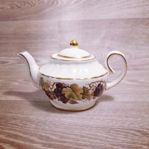 Puede incluir: Tetera de porcelana blanca con ribete dorado y pomo dorado en la tapa. La tetera presenta un diseño de uvas y hojas en tonos morados, verdes y marrones. El asa y el pico son curvos.