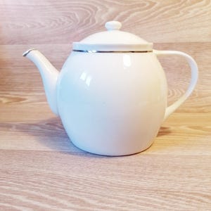 Può includere: Una teiera in ceramica bianca con corpo arrotondato, beccuccio curvo e manico ad anello. Il coperchio ha un piccolo pomello e un bordo argentato. La teiera è su una superficie di legno chiaro.