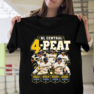 May include: A black t-shirt featuring a graphic of four baseball players in white uniforms with the text "NL CENTRAL 4-PEAT" at the top. Below the players are four yellow shield icons labeled "2023", "2024", "2025", and "2026 NL CENTRAL CHAMPIONS".