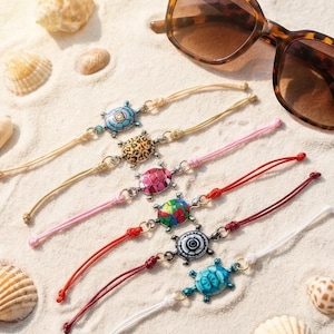 Könnte beinhalten: Eine Auswahl an Armbändern mit Schildkrötenanhängern und Kordeln in Beige, Rosa, Rot und Weiß. Eine Schildpatt-Sonnenbrille und Muscheln liegen auf sandigem Untergrund.