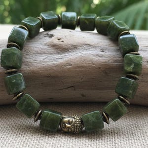 Puede incluir: Una pulsera de jade verde con cuentas en forma de cubo y detalles de latón antiguo. La pulsera presenta un pequeño dije de cabeza de Buda. Las cuentas están ensartadas con un cordón elástico. La pulsera se exhibe sobre un trozo de madera flotante.