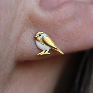 Puede incluir: Pendientes de pájaro de oro con detalles de esmalte blanco. El pájaro está posado en un pequeño poste.