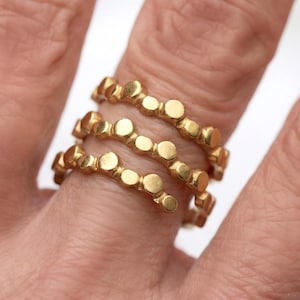 Könnte beinhalten: Drei goldfarbene Ringe, jeder mit einem sich wiederholenden Muster aus kleinen, erhabenen Kreisen. Die Ringe werden an einem Finger getragen und zeigen ihr spiralförmiges Design und ihren metallischen Glanz. Schmuck ist schlicht und elegant.