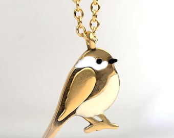 Puede incluir: Un collar con colgante de pájaro de color dorado. El pájaro tiene el pecho blanco y un ojo negro. La cadena del collar también es dorada. El pájaro está posado sobre una pata dorada.