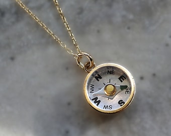 May include: A gold chain necklace with a small compass pendant. The compass face is silver with black markings for the cardinal directions: N, S, E, W, NE, NW, SE, SW. The compass needle is pointing towards the north.