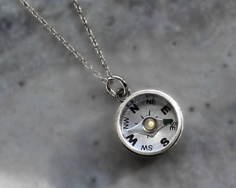 May include: A silver compass necklace with a small, round compass pendant. The compass face has black markings for the cardinal directions and a small, silver needle pointing north.