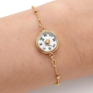 May include: Gold-coloured compass bracelet. The round compass face has black lettering indicating cardinal directions on a white background. The bracelet is attached to a delicate chain with small ball accents.