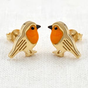 May include: A pair of gold-colored stud earrings shaped like robins. Each bird has an orange breast, black eye, and black beak. The earrings are on a white surface.