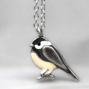 Puede incluir: Un collar de plata con un peque&ntilde;o colgante de p&aacute;jaro detallado. El p&aacute;jaro es blanco con detalles negros y una cadena de plata.