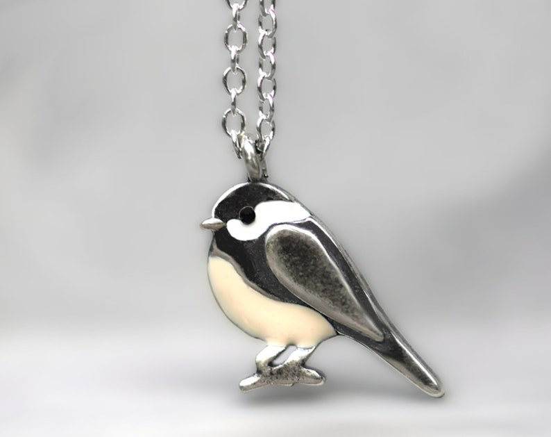 Puede incluir: Un collar de plata con un peque&ntilde;o colgante de p&aacute;jaro detallado. El p&aacute;jaro es blanco con detalles negros y una cadena de plata.