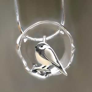 Puede incluir: Un collar de plata con un colgante que presenta un peque&ntilde;o p&aacute;jaro posado en una rama dentro de un c&iacute;rculo. El p&aacute;jaro es blanco con detalles negros.