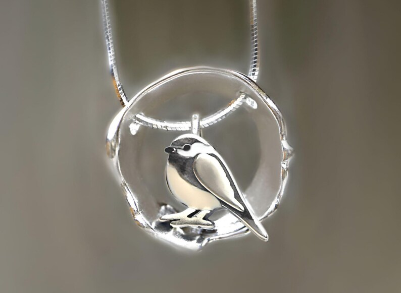 Puede incluir: Un collar de plata con un colgante que presenta un peque&ntilde;o p&aacute;jaro posado en una rama dentro de un c&iacute;rculo. El p&aacute;jaro es blanco con detalles negros.