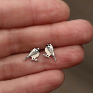 Puede incluir: Un par de pendientes de bot&oacute;n de plata en forma de p&aacute;jaro. Los p&aacute;jaros miran hacia la derecha y tienen detalles blancos y grises.