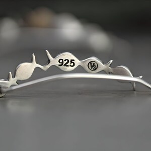 May include: Silver ear cuff with a curved design featuring a series of small fish shapes. The cuff is marked with "925" and "VS" on the back.