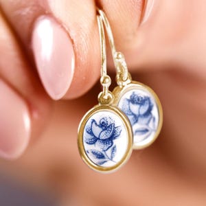 Puede incluir: Pendientes dorados con colgantes ovalados. Cada colgante presenta un diseño de rosa azul sobre fondo blanco, enmarcado por un borde dorado. Los pendientes están sostenidos por los dedos de una persona.