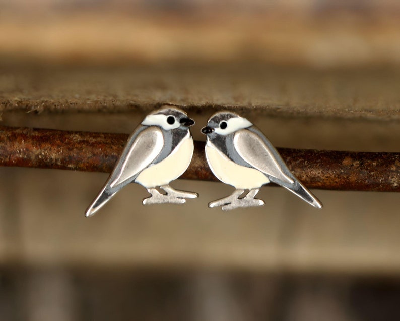 Puede incluir: Un par de pendientes plateados con un dise&ntilde;o de p&aacute;jaro peque&ntilde;o. Los p&aacute;jaros son blancos con detalles grises y tienen ojos negros.