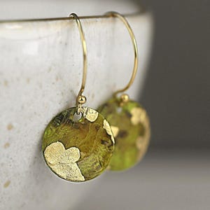 May include: A pair of gold-colored earrings with a circular, textured design. The earrings have a green and gold color scheme, with gold leaf accents. The earrings are hanging from a gold-colored hook.
