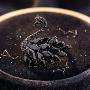 May include: A black swan brooch with a detailed design. The swan's body is composed of black, teardrop-shaped stones, and the neck and head are adorned with smaller black stones. The brooch is set against a dark, textured background.