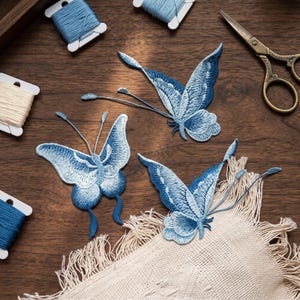 May include: Three blue embroidered butterfly patches with detailed wing patterns, on a wooden surface. Spools of thread and gold scissors are also visible. A piece of beige fabric with fringe is also present, suggesting a craft theme.