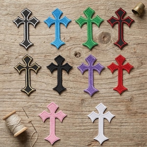 Op de afbeelding: Een verzameling geborduurde kruisvormige patches in diverse kleuren, waaronder zwart, blauw, groen, rood, goud, paars, roze en wit. De patches zijn gerangschikt op een houten oppervlak, met een garenklos en een vingerhoed.