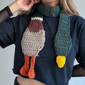 Puede incluir: Bufanda de ganchillo hecha a mano con forma de pato. La bufanda tiene cuello y cola verdes, cuerpo marrón y beige y patas naranjas. El pico del pato es amarillo. La bufanda se lleva alrededor del cuello.