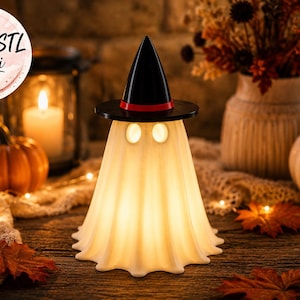 May include: A white, glowing ghost-shaped lamp wearing a black witch hat with a red band. The ghost has two circular eye cutouts and a smooth, draped fabric-like texture. A circular badge in the top left corner displays the text 3MF & STL Datei.