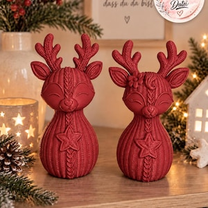 May include: Two dark red reindeer figurines with a textured knit pattern. Each deer features closed eyes, a heart-shaped nose, and a prominent star on its chest. The antlers have a cable-knit design, and one deer has a small flower on its head.