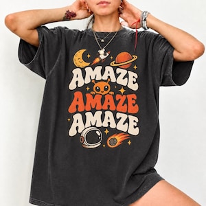 May include: A charcoal grey short-sleeve t-shirt featuring a space-themed graphic with the text "AMAZE" repeated three times. The design includes a crescent moon, a rocket, a planet, an alien face, an astronaut helmet, and a comet in orange.