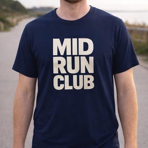 May include: Navy blue t-shirt with the text "MID RUN CLUB" in large, white, block letters. The shirt is a crew neck style and appears to be made of a soft material. The image is taken outdoors.