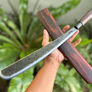 以下が含まれることがあります： 手作業で鍛造されたマチェットで、質感のあるダークグレーの金属製ブレードと滑らかなダークブラウンの木製ハンドルを備えています。ブレードの柄の近くには刻印されたロゴがあります。赤と黒の木目模様が特徴的な、お揃いの木製鞘が付属しています。