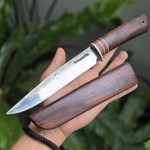 May include: A fixed-blade knife with a polished steel blade featuring a textured spine and the text fonsaenha. The handle is crafted from dark brown wood with two silver-coloured rings near the bolster. A matching wooden sheath is included.
