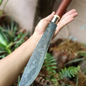 May include: A fixed-blade knife featuring a long, tapered steel blade with a dark, mottled finish. The handle is crafted from smooth, reddish-brown wood, connected to the blade by a polished silver-coloured metal bolster and a thin white ring.