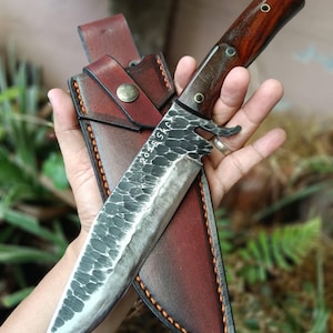 May include: A fixed-blade knife with a dark, hammered metal finish on the blade, featuring the engraved text 2025 SKF. The handle is made of polished dark wood with three brass rivets. It is paired with a brown leather sheath with orange stitching.