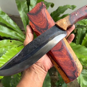 May include: A handcrafted knife with a dark blade and a wooden handle and sheath. The handle and sheath feature a reddish-brown wood grain pattern. The blade has a series of star-shaped designs and the word "SALU" etched into it.
