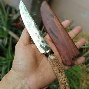 Pode incluir: Uma faca de lâmina fixa com uma lâmina de metal texturizada e manchada, um espaçador de latão e preto, e um cabo de chifre curvo de cor castanho-claro. A faca é acompanhada por uma bainha de madeira lisa castanho-escura com veios visíveis.