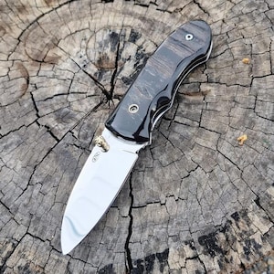 May include: A folding pocket knife with a polished steel blade and a handle featuring dark wood and black accents. The knife has a brass-colored pivot and a curved design. The blade is open, displaying its sharp edge.