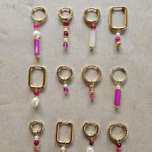 Könnte beinhalten: Eine Sammlung goldfarbener Ohrringe mit verschiedenen Designs. Einige Ohrringe haben rosa und weiße Perlen, während andere rosa rechteckige oder weiße rechteckige Anhänger haben. Die Ohrringe sind auf einer neutral getönten Oberfläche angeordnet.