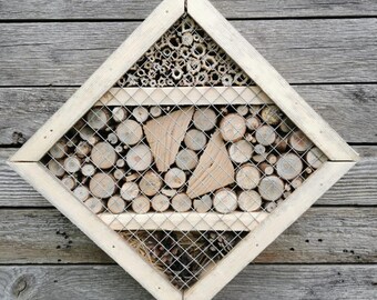 Könnte beinhalten: Ein rautenförmiges Insektenhotel aus Holz mit hellem Rahmen. Das Haus ist in Abschnitte unterteilt, die mit Bambusröhren, kleinen Holzscheiben und Holzstücken gefüllt sind, alle hinter Drahtgeflecht. An einer verwitterten Holzwand montiert.