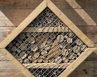 Puede incluir: Un hotel para insectos de madera en forma de rombo con un marco marrón claro y una parte frontal de malla metálica. El interior está relleno de diversos materiales naturales, como cañas huecas, troncos perforados y piñas.
