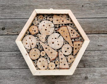 Könnte beinhalten: Sechseckiges Bienenhaus aus Holz mit hellem Holzrahmen und Drahtgitterfront. Das Innere ist mit Holzstücken in verschiedenen Größen und Formen gefüllt, die Lebensräume für Bienen bieten.