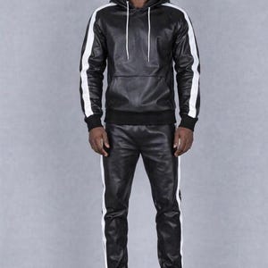 K&ouml;nnte beinhalten: Schwarzer Leder-Hoodie und passende Hose mit wei&szlig;en Streifen an &Auml;rmeln und Seiten. Der Hoodie hat einen Kordelzug und eine Vordertasche. Die Hose hat einen elastischen Bund und B&uuml;ndchen. Dazu wei&szlig;e Sneaker.