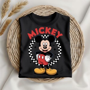 May include: A black t-shirt featuring a graphic of Mickey Mouse standing with hands on hips. The design includes the word MICKEY in red letters above a black and white checkered circle background with small star accents and yellow shoes.