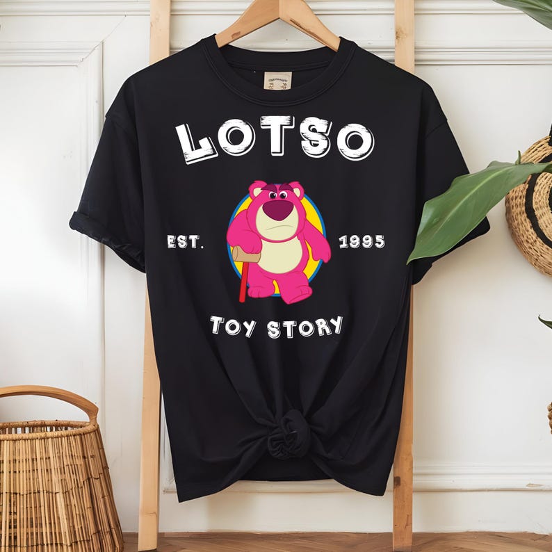 Puede incluir: Camiseta negra con la palabra "LOTSO" sobre un dibujo de un oso rosa sosteniendo un mazo, dentro de un c&iacute;rculo amarillo. El texto "EST. 1995" y "TOY STORY" tambi&eacute;n est&aacute;n impresos en la camiseta.