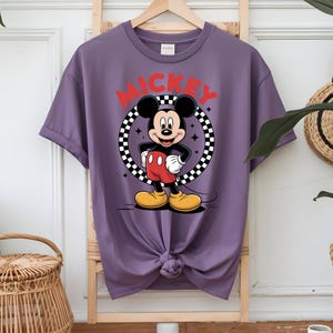 May include: A purple short-sleeve t-shirt featuring a graphic of Mickey Mouse standing inside a black and white checkered circle. The word MICKEY is printed in large red capital letters above the character. The shirt has a crew neckline.